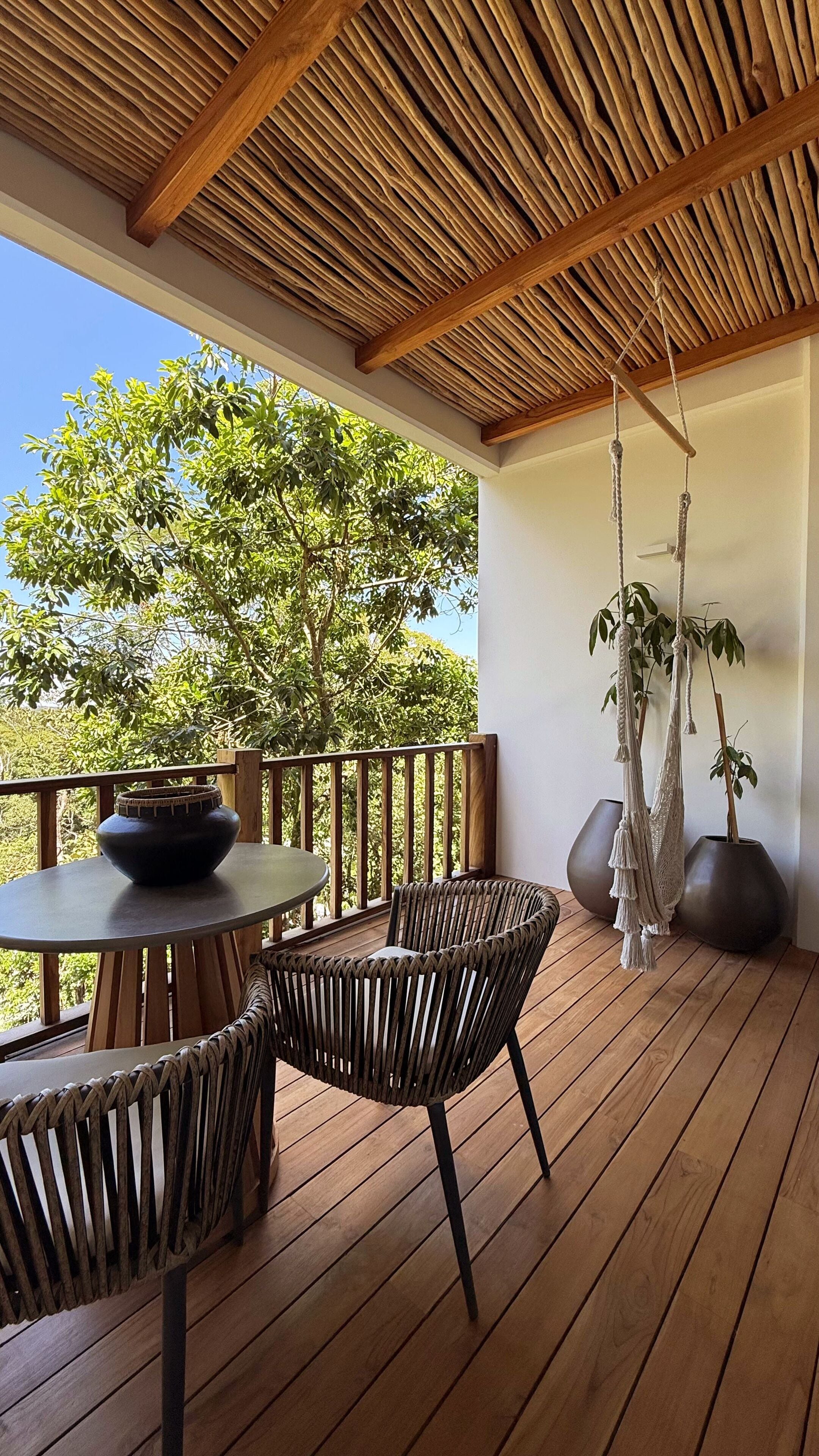 A wooden deck with a round table and chairs, a hanging macrame plant, and a view of trees.