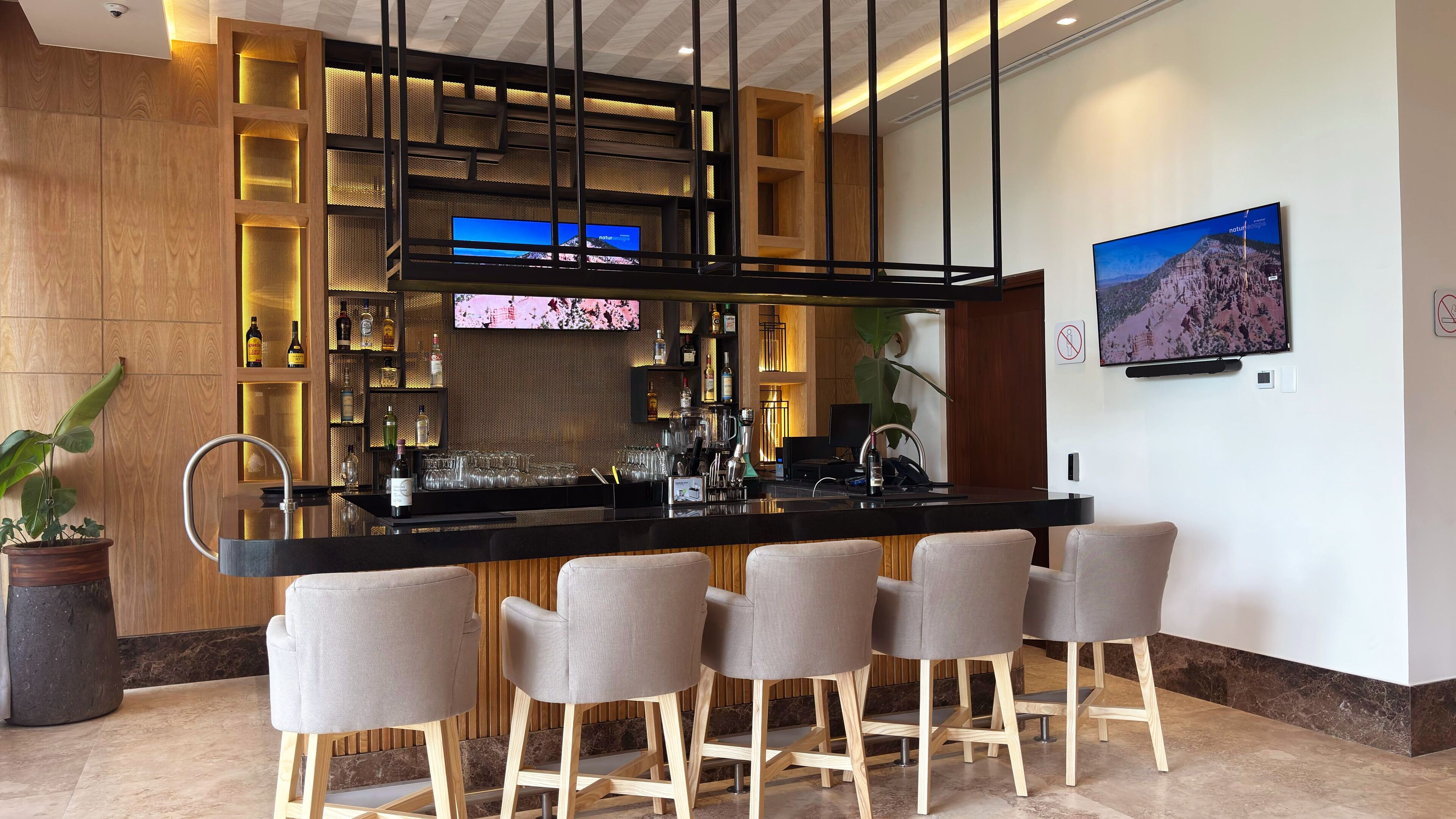 A modern bar area with a black countertop, four grey bar stools, and a television mounted on the wall displaying a mountain scene.