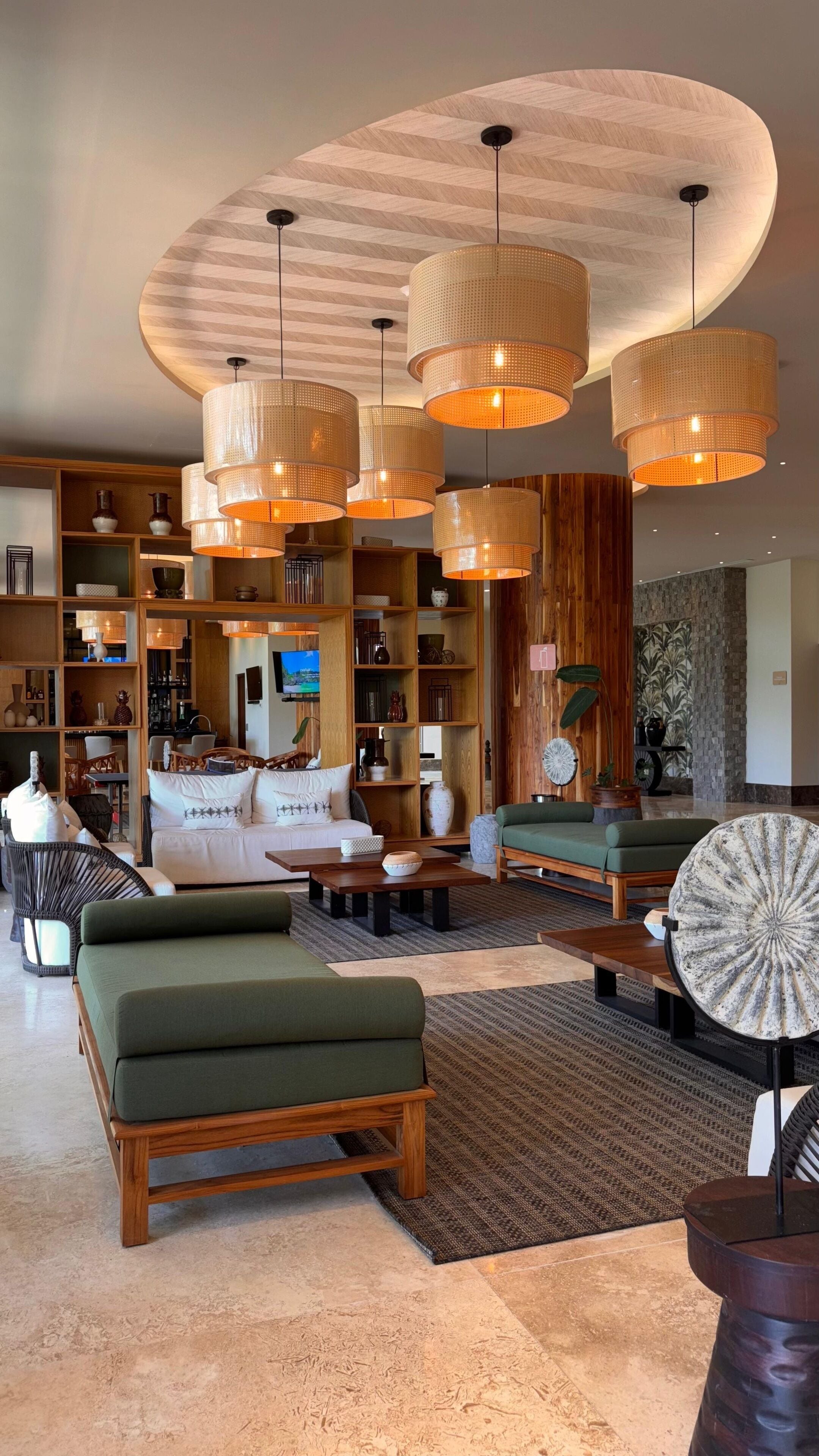 A spacious living area with multiple seating options, including sofas and a coffee table. There are several bookshelves filled with books and decorative items. The ceiling features a unique wooden design with hanging pendant lights.
