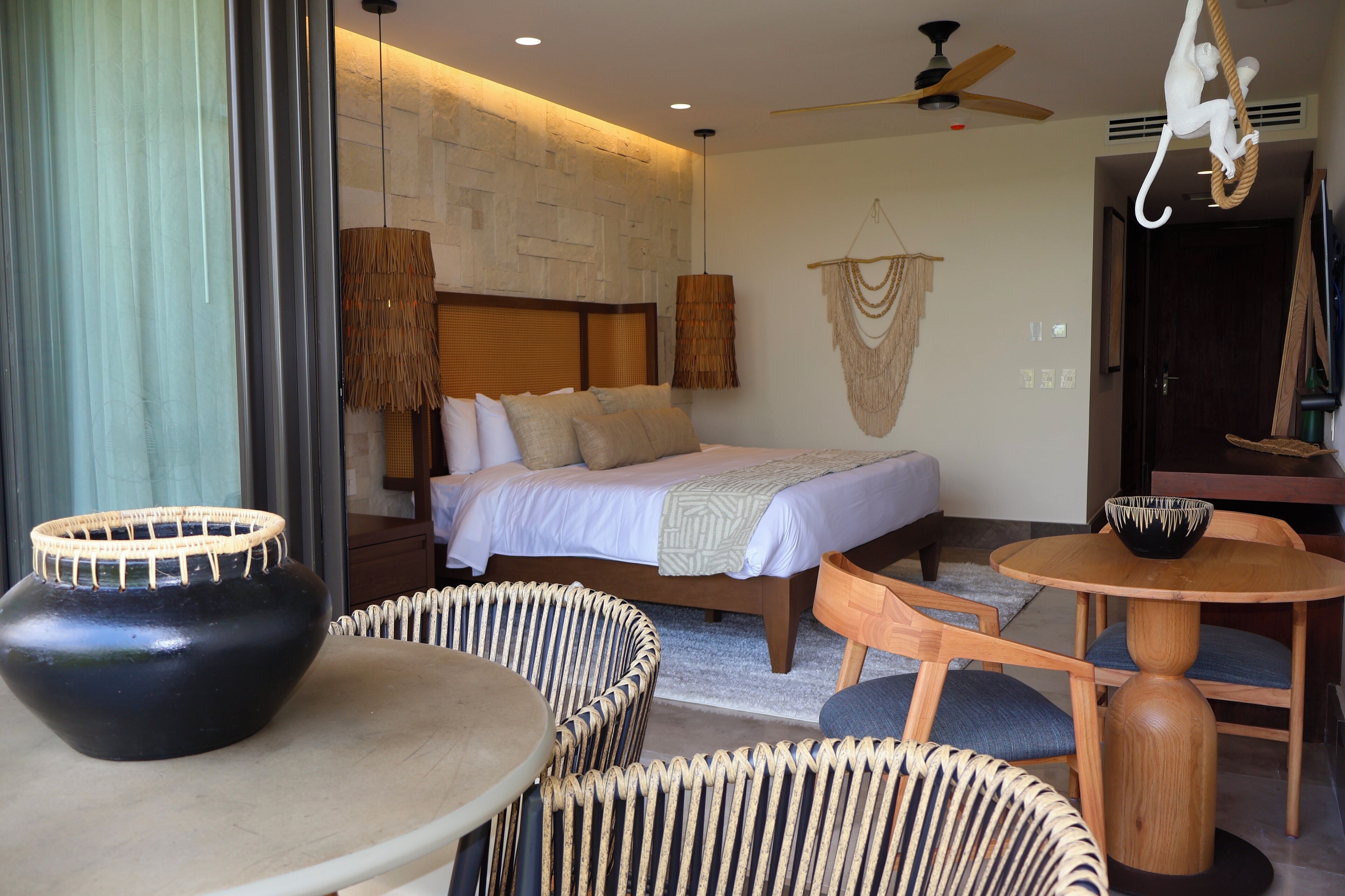 A modern hotel room with a bed, wicker furniture, a round wooden table, and a ceiling fan.
