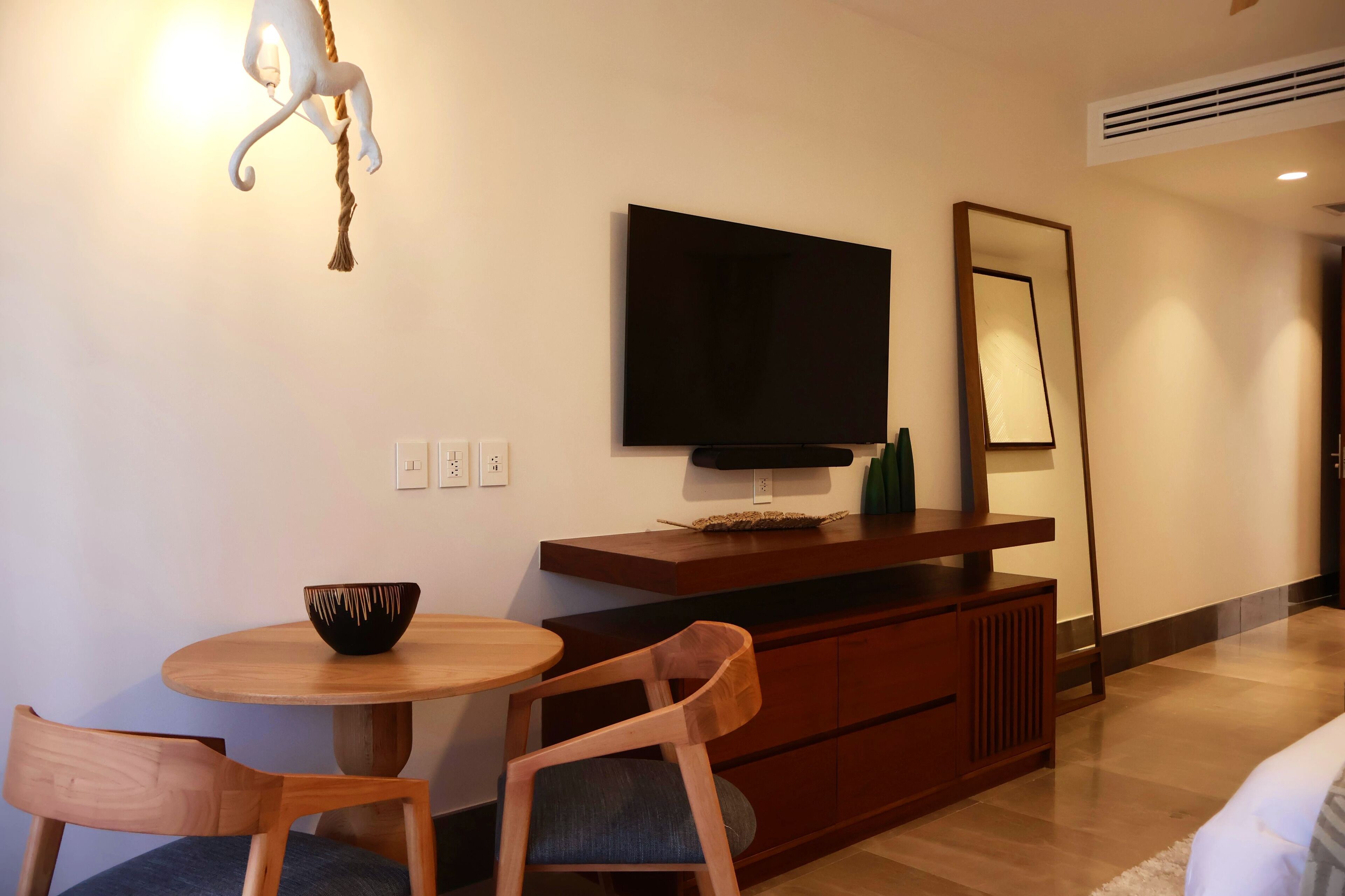 A modern hotel room with a wooden dining table, two chairs, a flat-screen TV mounted on the wall, a wooden cabinet, and a large mirror.