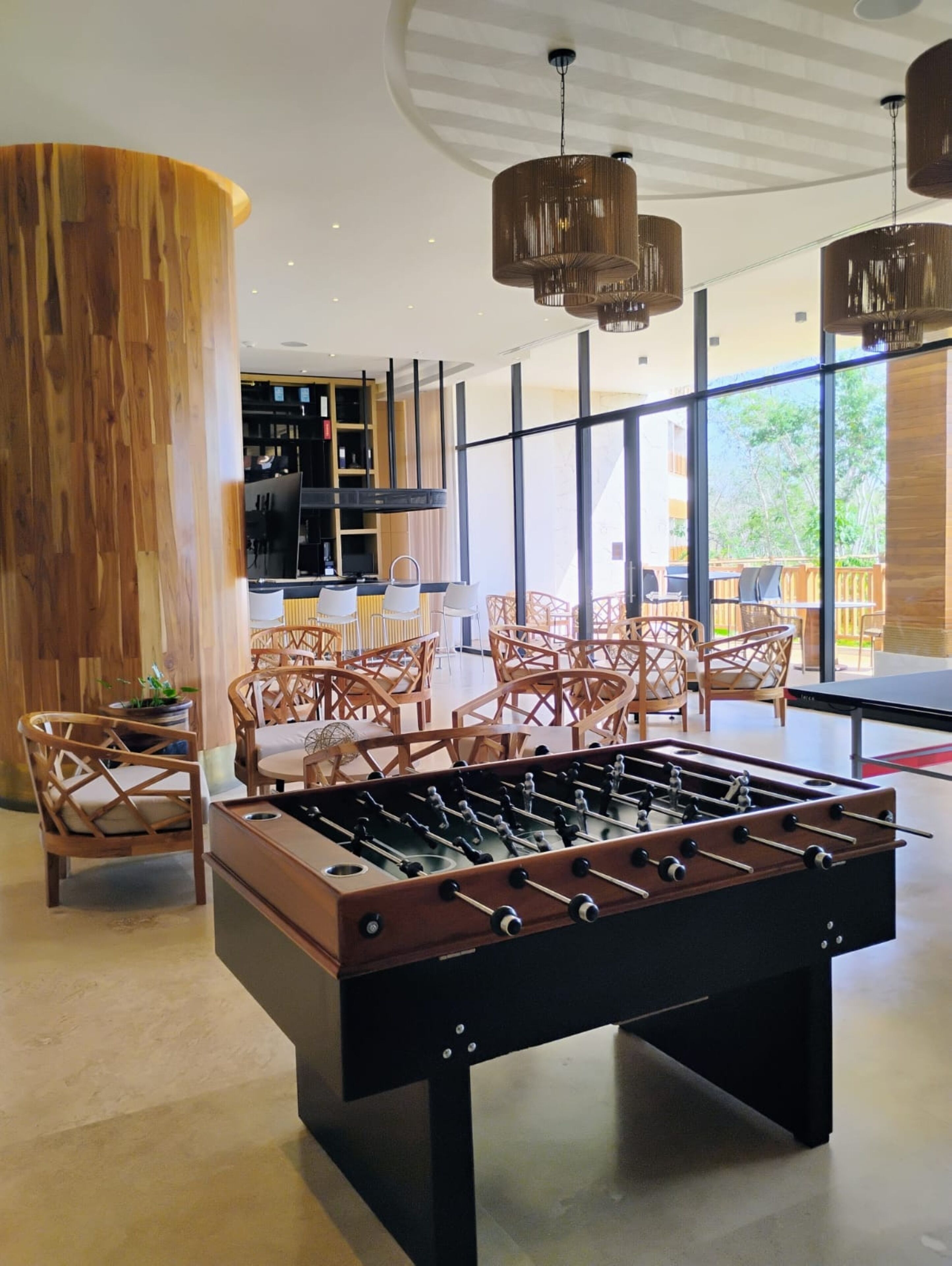 Un espacio interior moderno con mesa de fútbol de mesa, sillas de mimbre y una zona de bar con un ventanal que ofrece vistas al exterior.