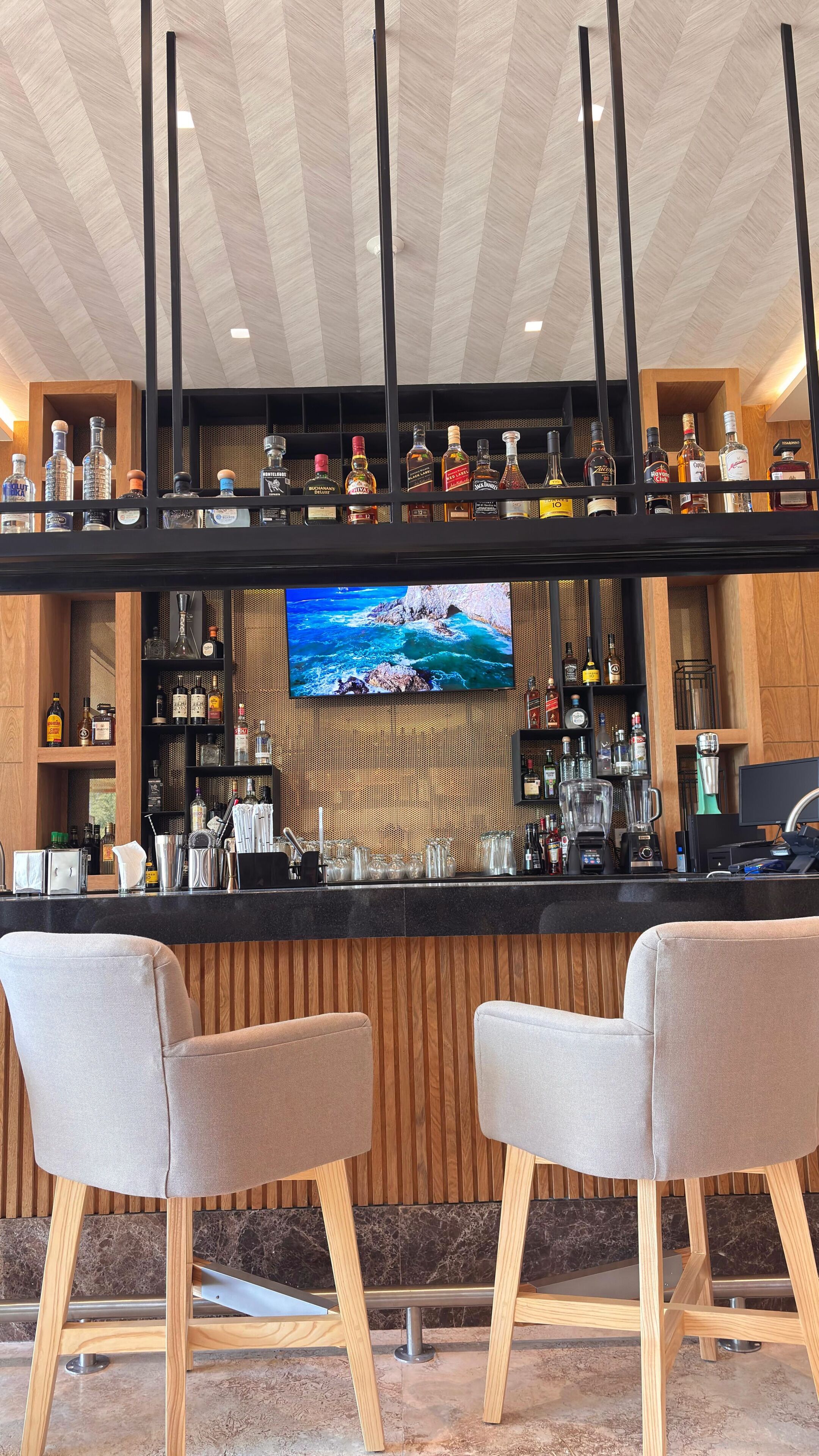 A bar area with two cushioned bar stools, a television mounted on the wall, and shelves stocked with various liquor bottles.