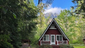 Une petite cabane rouge, dotée d’un porche et d’un escalier menant à la porte, entourée d’une forêt dense et avec une montagne en arrière-plan.