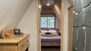 Une chambre avec un plafond en pente, un lit recouvert d’une couverture à carreaux, une fenêtre donnant sur les arbres, une commode en bois et un mur en pierre.