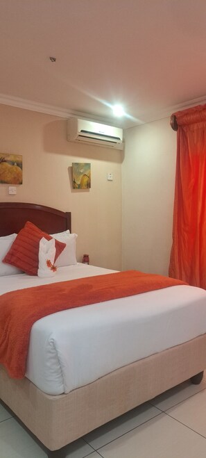 Een net opgemaakt bed met een wit-oranje deken, een houten hoofdbord en twee aan de muur bevestigde schilderijen.