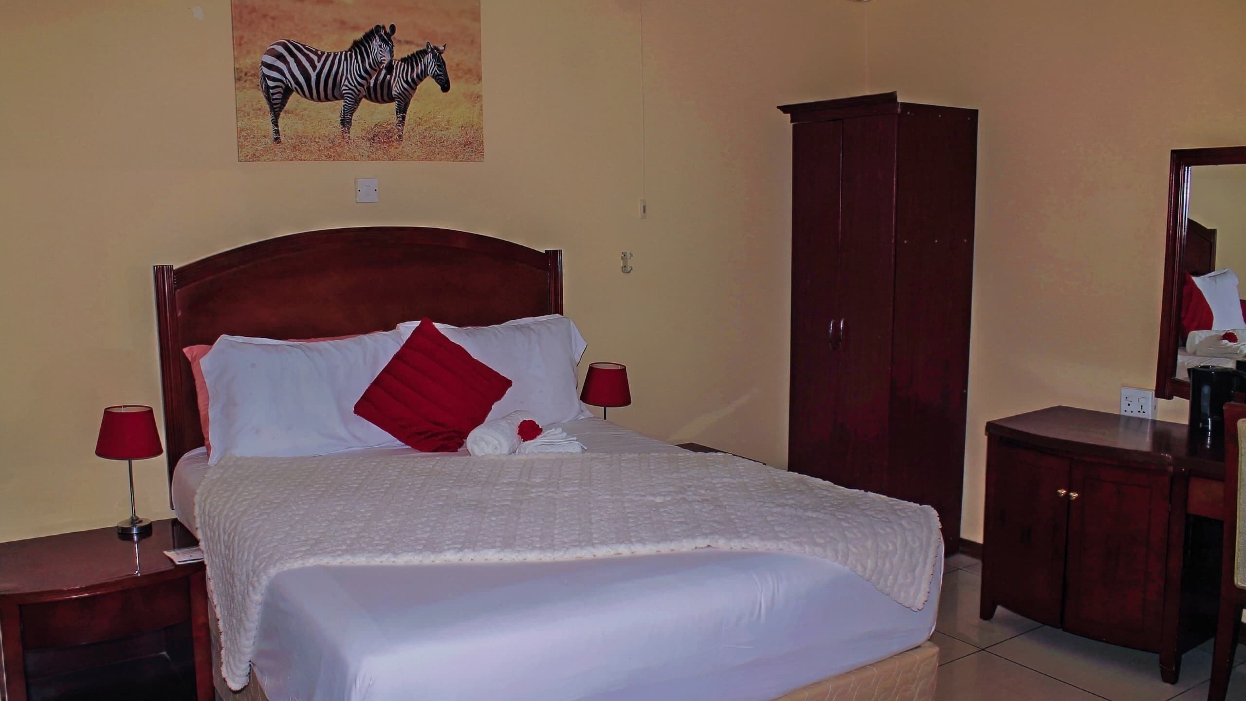 Una cama bien hecha con sábanas blancas y almohadas rojas, un armario de madera, una lámpara roja y un cuadro de cebra en la pared.