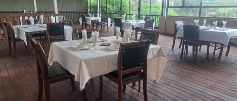 Un espacio de comedor al aire libre con mesas dispuestas para el servicio, rodeado de sillas de madera y cubiertas con manteles blancos.
