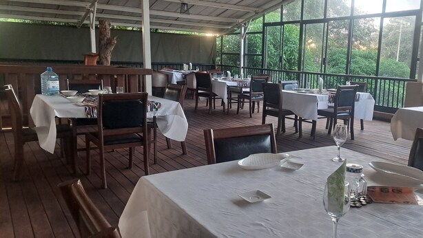 A restaurant with multiple tables and chairs, covered by a roof, and large windows offering a view of greenery outside. - Comfort Palace  (Kasane)