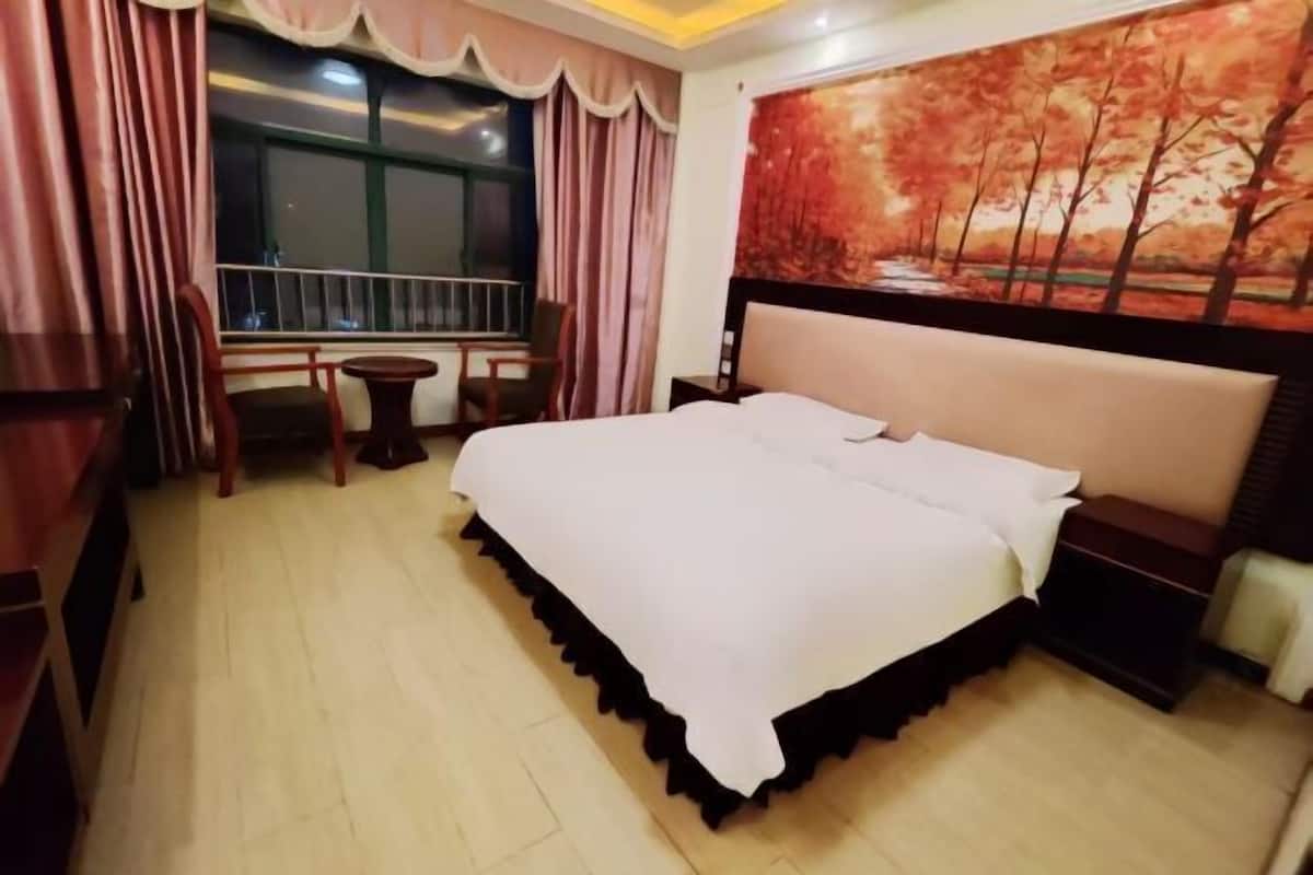 A hotel room with a bed, a small table, a chair, a window with curtains, and a painting on the wall.