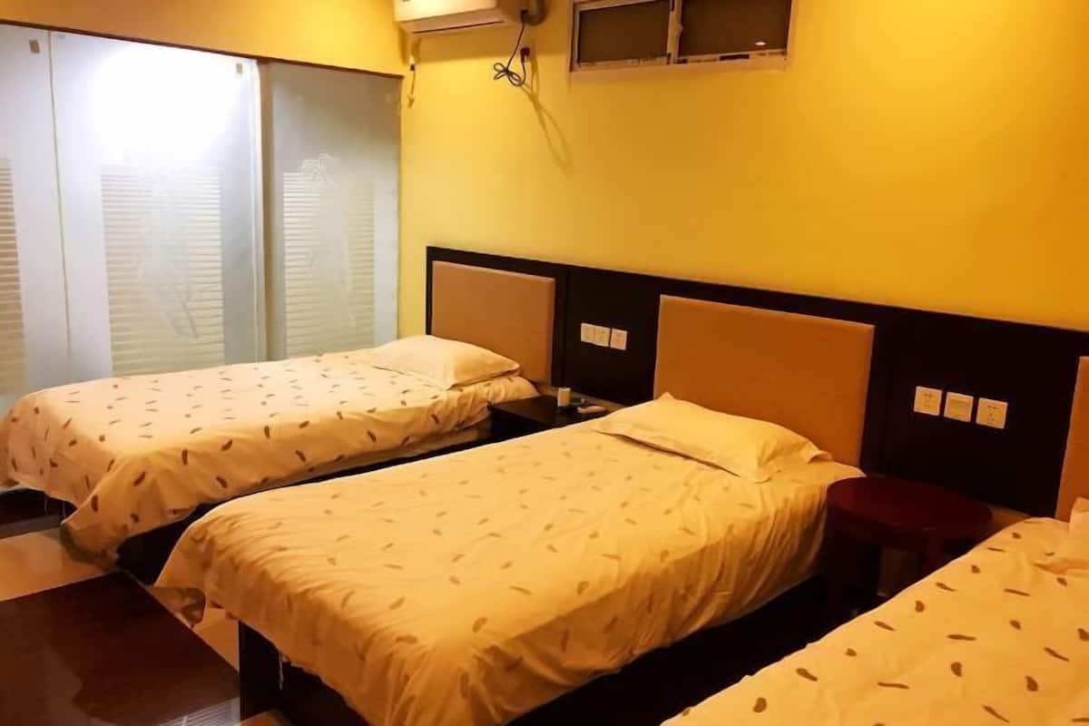A hotel room with two beds, a small table, a wall-mounted air conditioner, and a window with blinds.