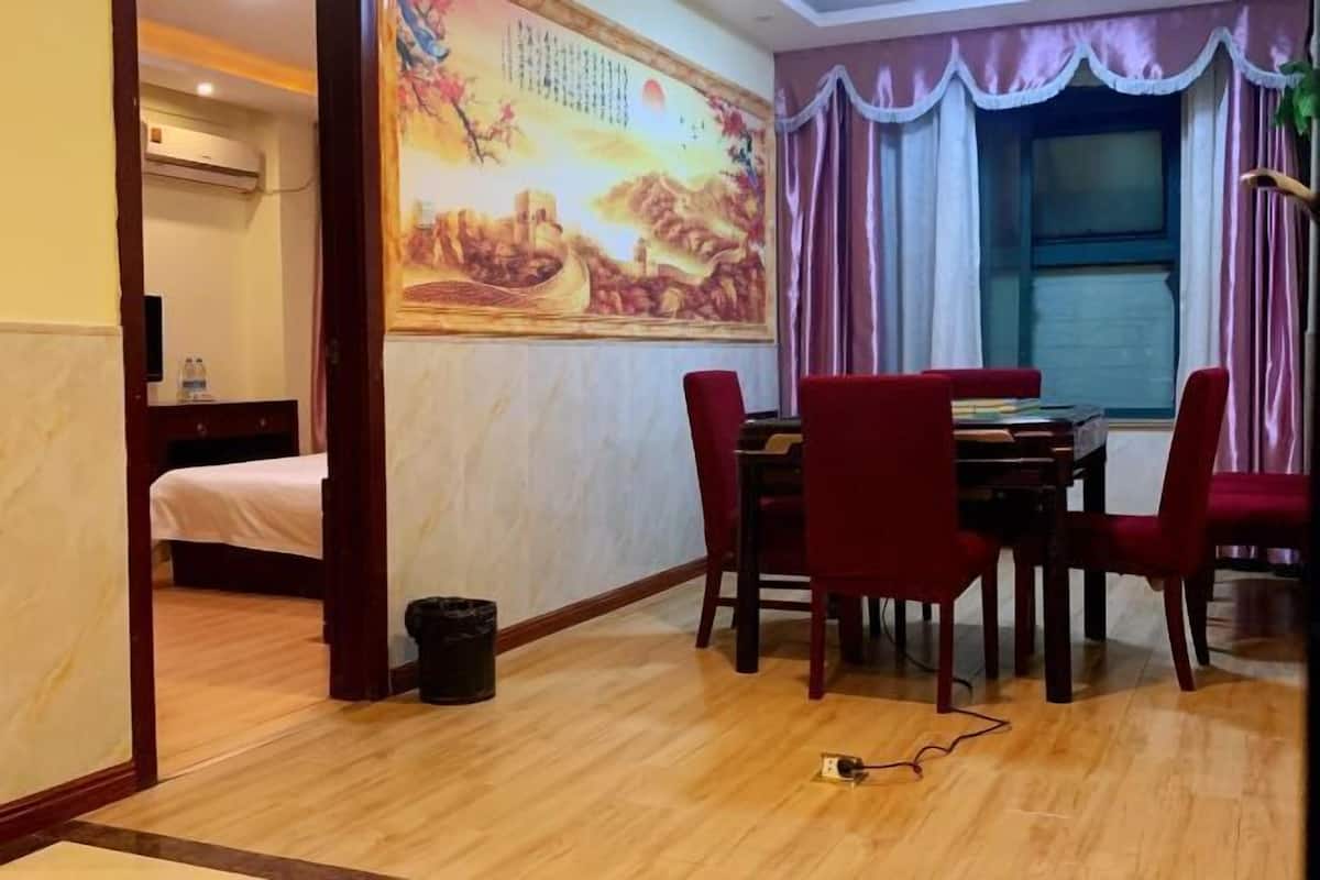 A hotel room with a wooden floor, a dining table with red chairs, a bed with white bedding, a wall mural, and a window with curtains.