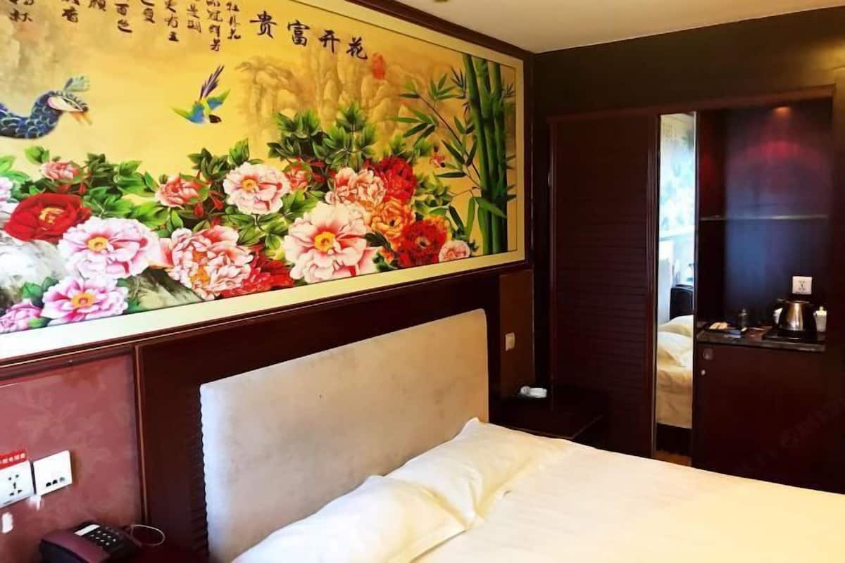 A hotel room with a bed, a wall-mounted painting, a wooden headboard, a bedside table, and a nightstand.