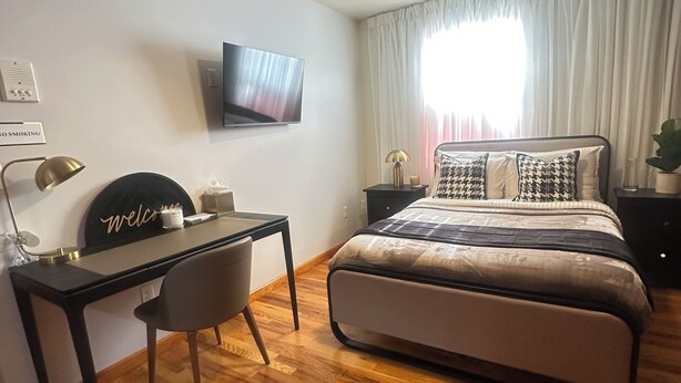 A bedroom with a bed, a desk, a chair, a TV, a ceiling fan, and a window with curtains. - Amelia Suites Newark – Cozy stay with AC, Wi-Fi & NYC access! (Newark)