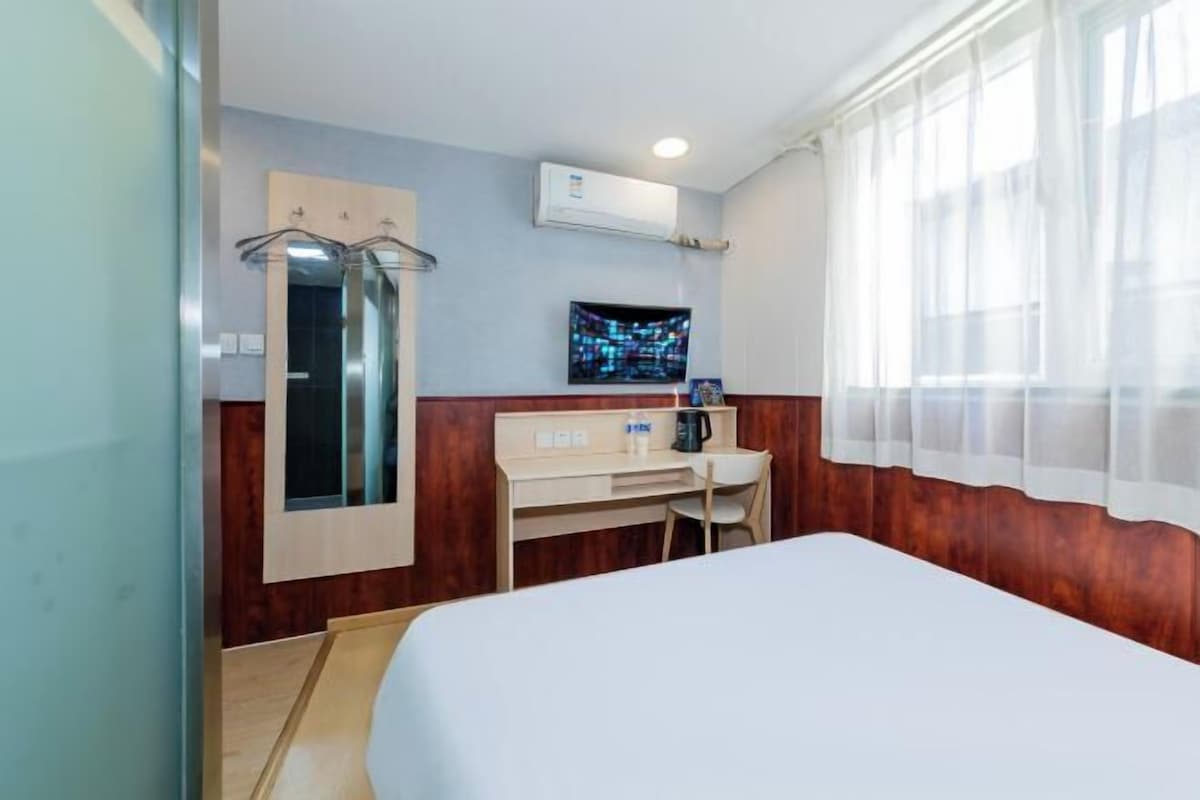 A hotel room with a bed, a desk, a chair, an air conditioning unit, a television, and a window with sheer curtains.