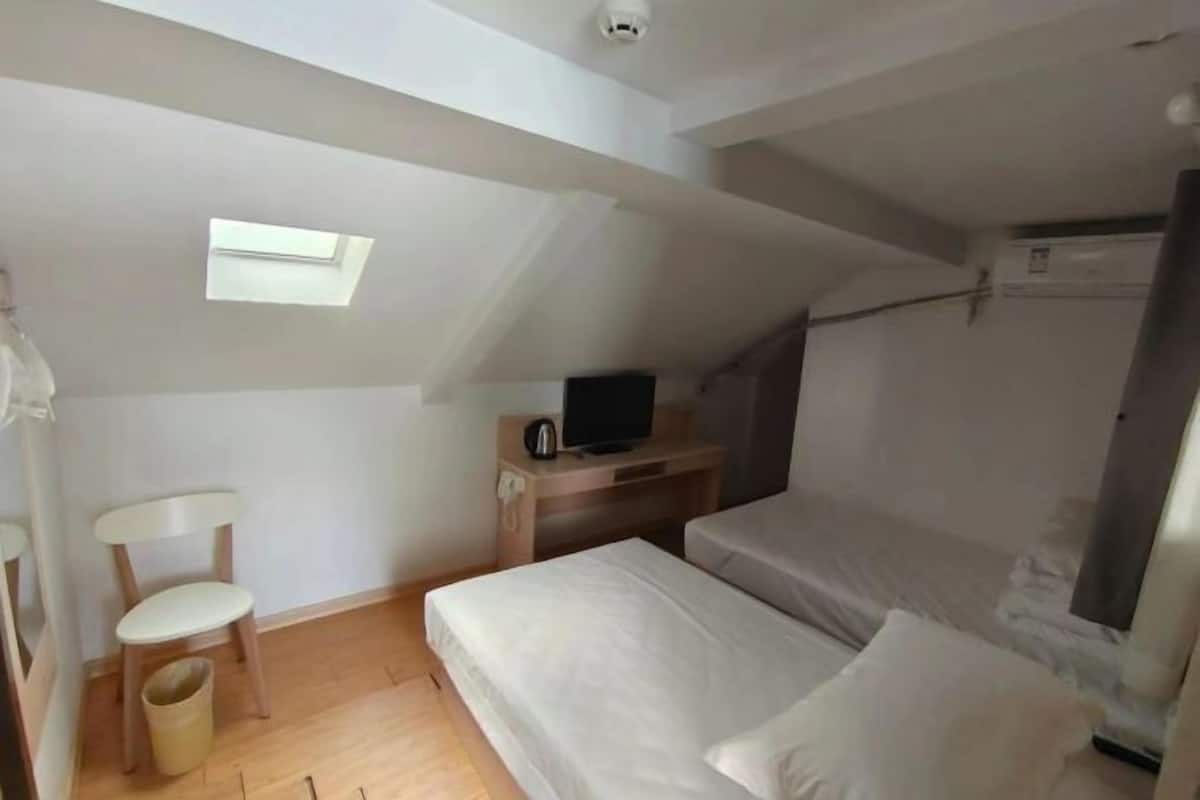 Um apartamento pequeno, de um único quarto, com teto inclinado, janela, escrivaninha com televisão, cadeira e cama.