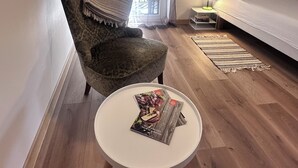 Ein Zimmer mit einem Bett, einem Sessel, einem kleinen Tisch mit Zeitschriften, einer Lampe und Blick durch ein Fenster.