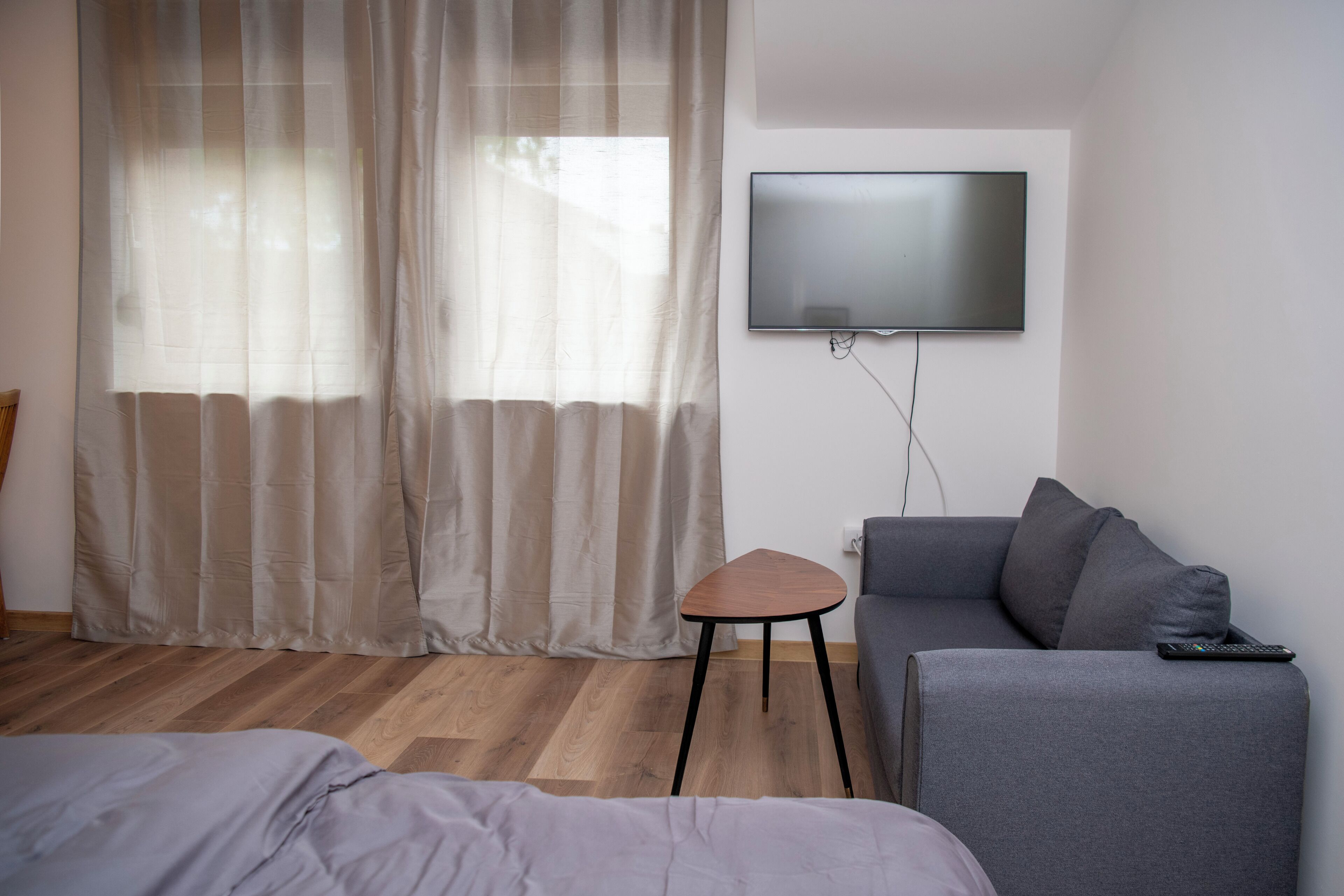 A modern hotel room with a bed, a gray sofa, a small wooden stool, a TV mounted on the wall, and sheer curtains.