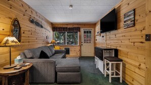 Un intérieur de chalet chaleureux avec un canapé d’angle, un téléviseur et un mur en bois sur lequel sont affichées les règles de la maison.