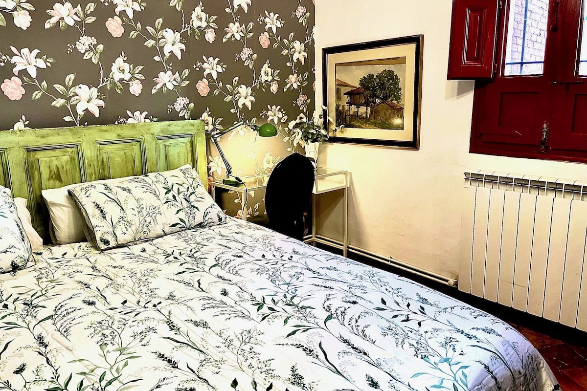 Een slaapkamer met bloemenbehang, een groen hoofdeinde, een ingelijst schilderij aan de muur en een rode deur.