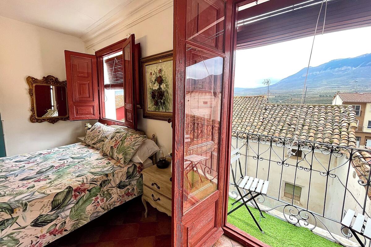 Een slaapkamer met een bed, een dressoir, een spiegel en een raam met uitzicht op een pannendak en verre bergen.