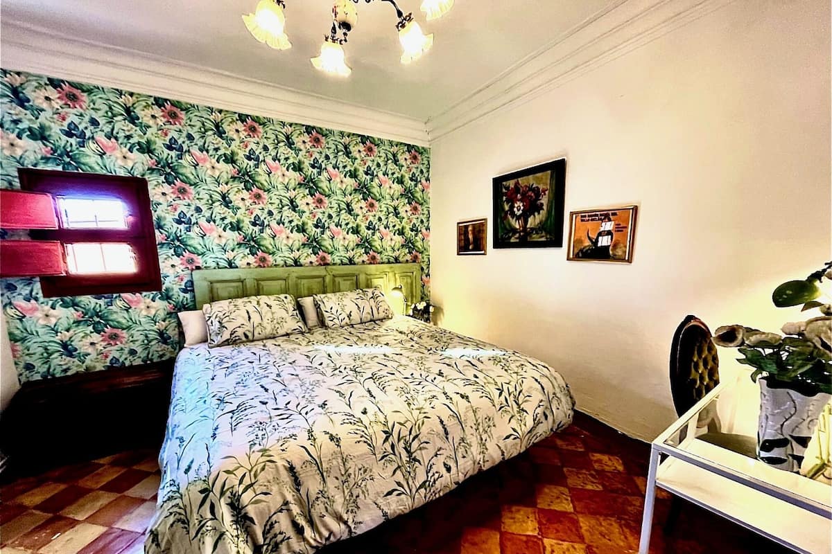 Een slaapkamer met bloemenbehang, een bed met een beddekleed met dessin, een nachtkastje, een lamp en een ingelijste foto aan de muur.