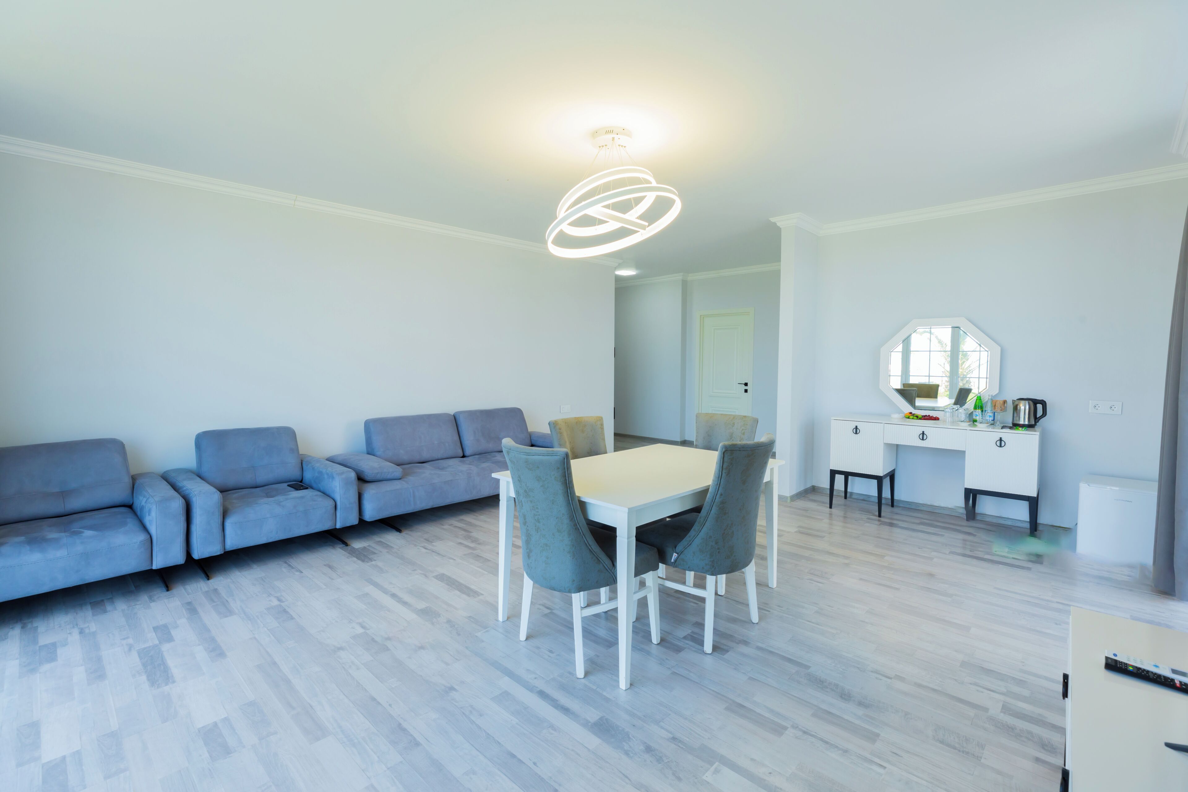 A modern living room with a sofa set, a dining table with chairs, a kitchen area with a countertop and appliances, and a large mirror.