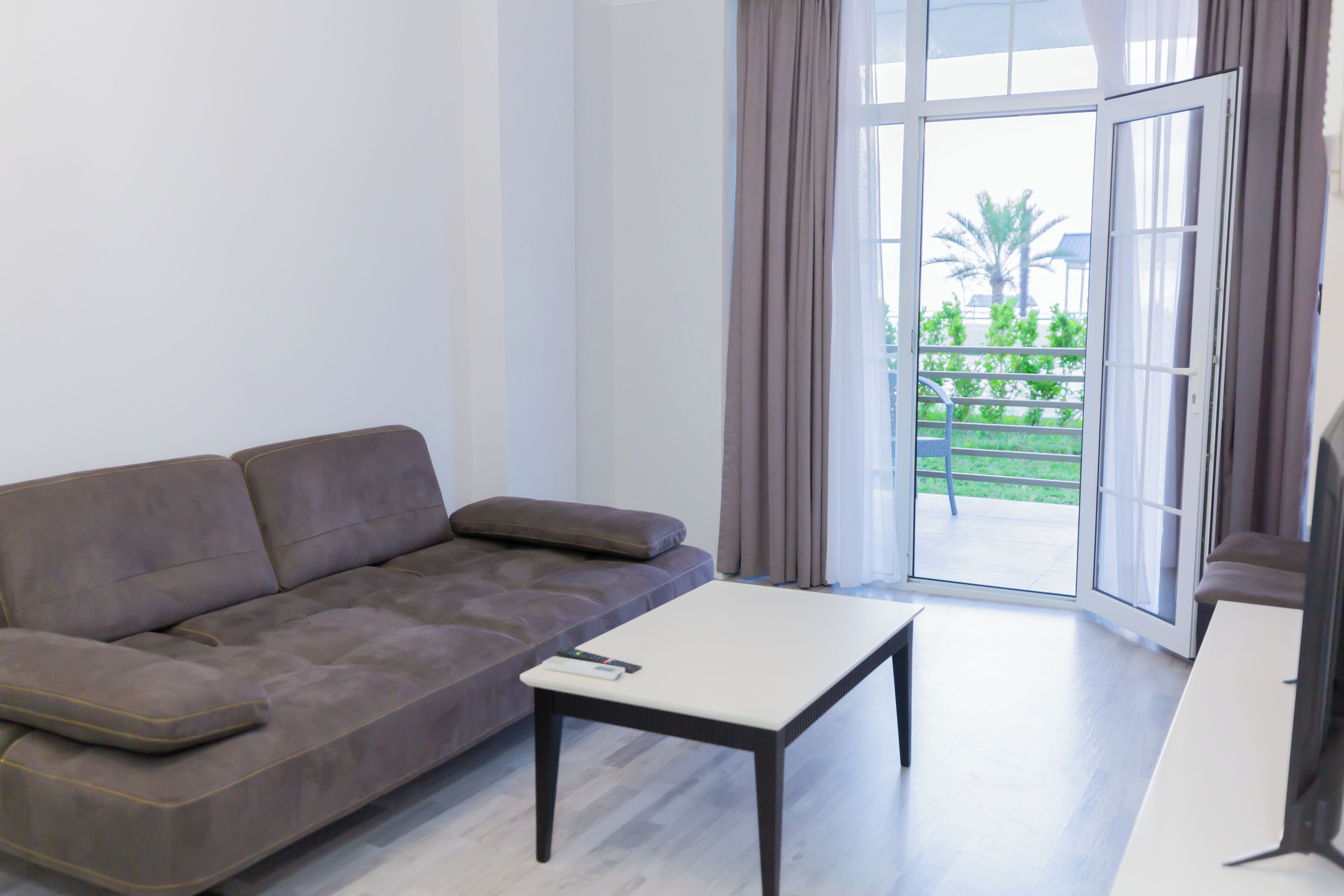 A modern living room with a grey sofa, a white coffee table, and a sliding glass door leading to a balcony with a view of greenery.