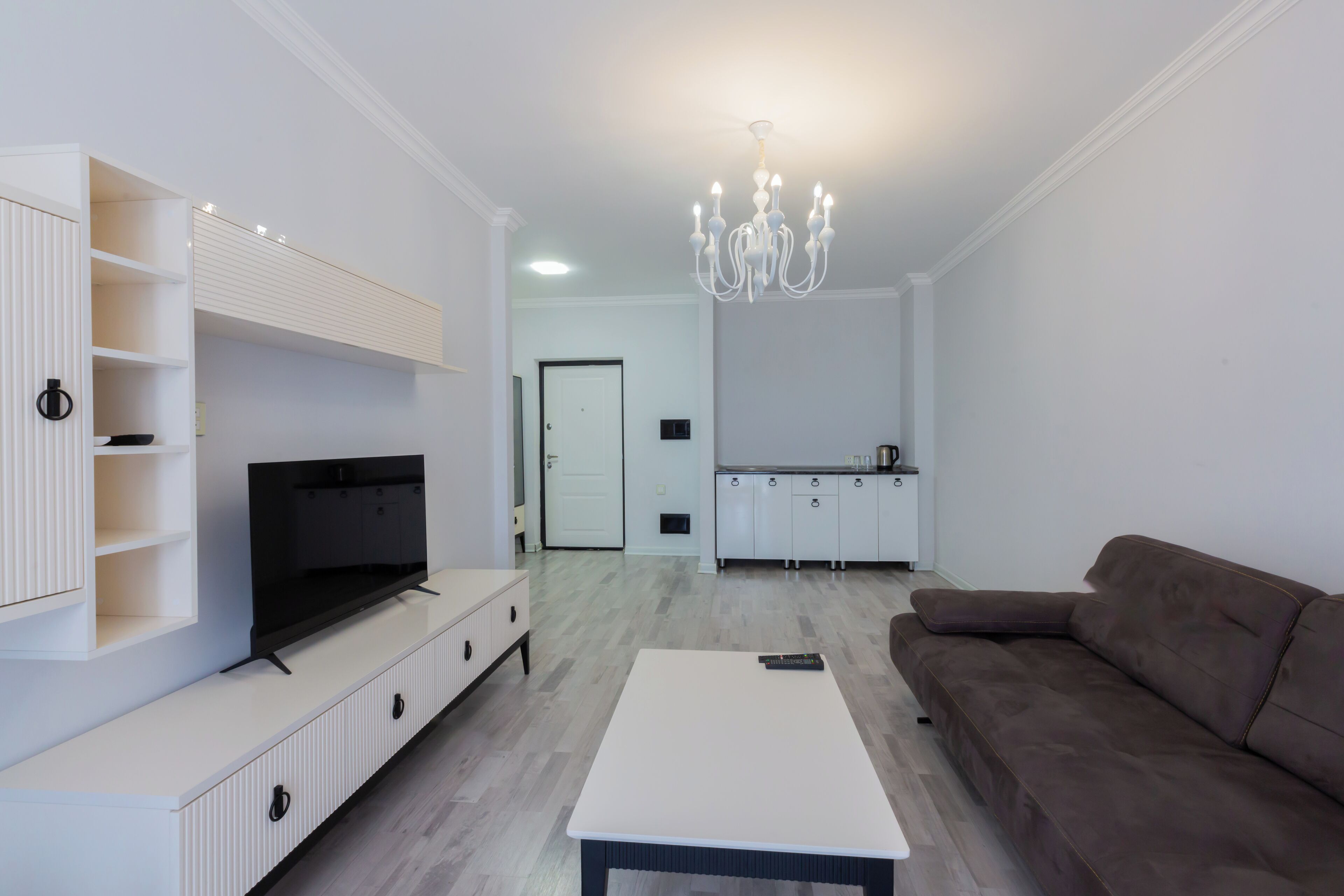 Un salon moderne avec un téléviseur à écran plat, un canapé marron, une table basse blanche et une cuisine en arrière-plan.