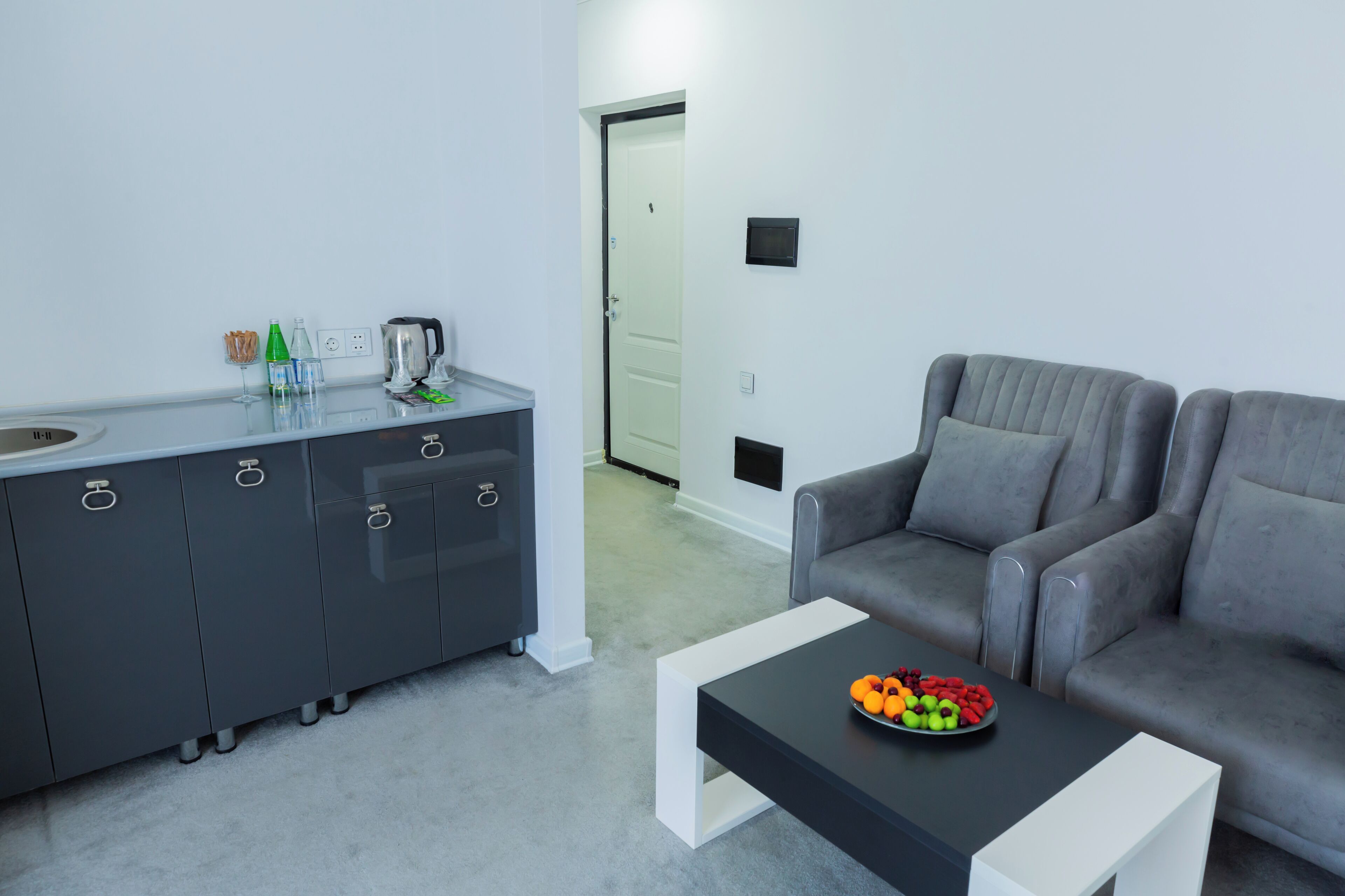 Een moderne woonkamer met een grijze bank, een bijzettafeltje met fruit en een grijs kastje met keukenapparatuur.