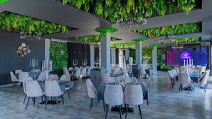 A restaurant with multiple dining tables and chairs, green foliage on the ceiling, chandeliers, and a neon sign in the background.