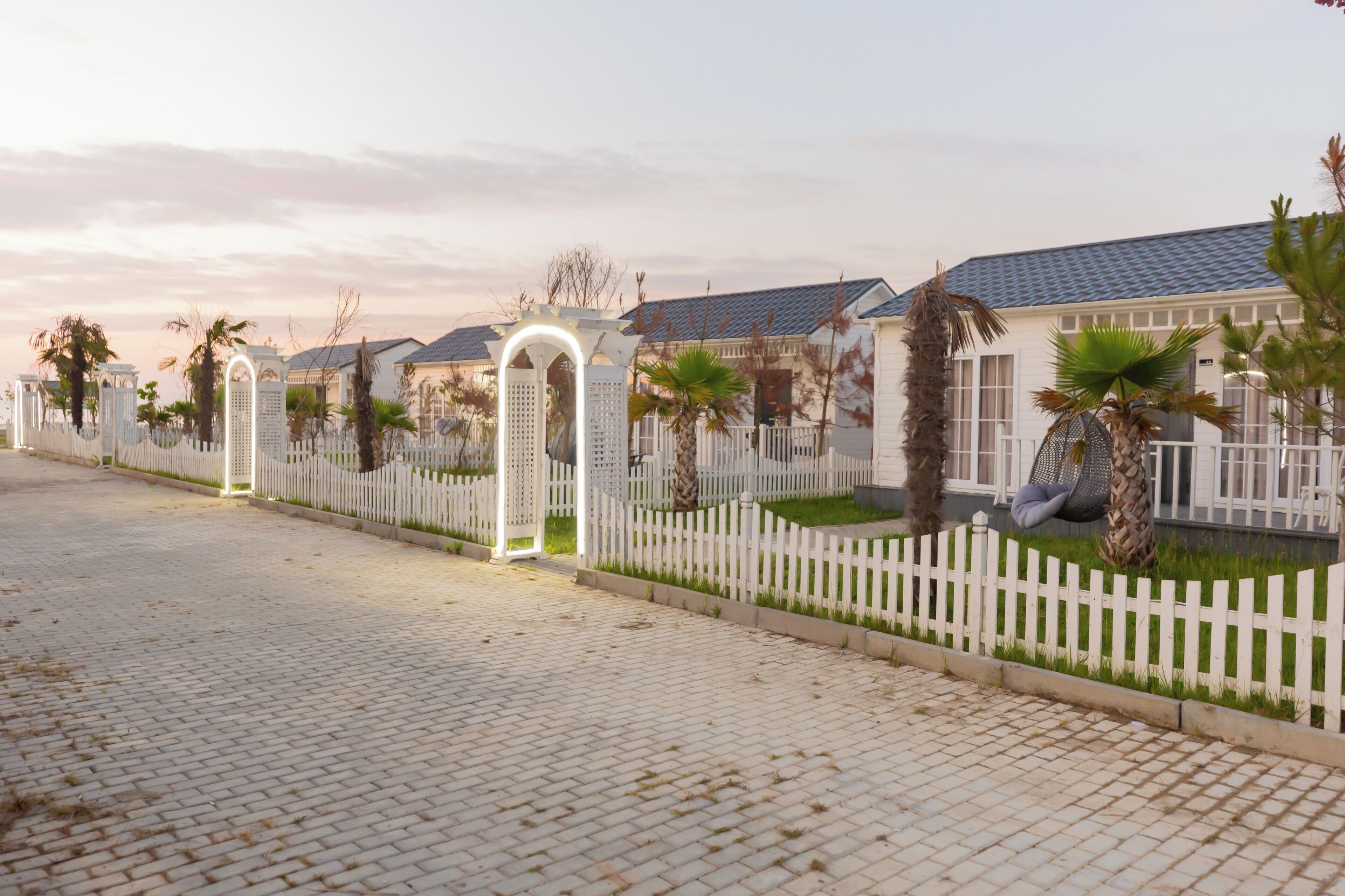 A paved pathway with a white fence and palm trees, leading to a row of houses with a blue roof.