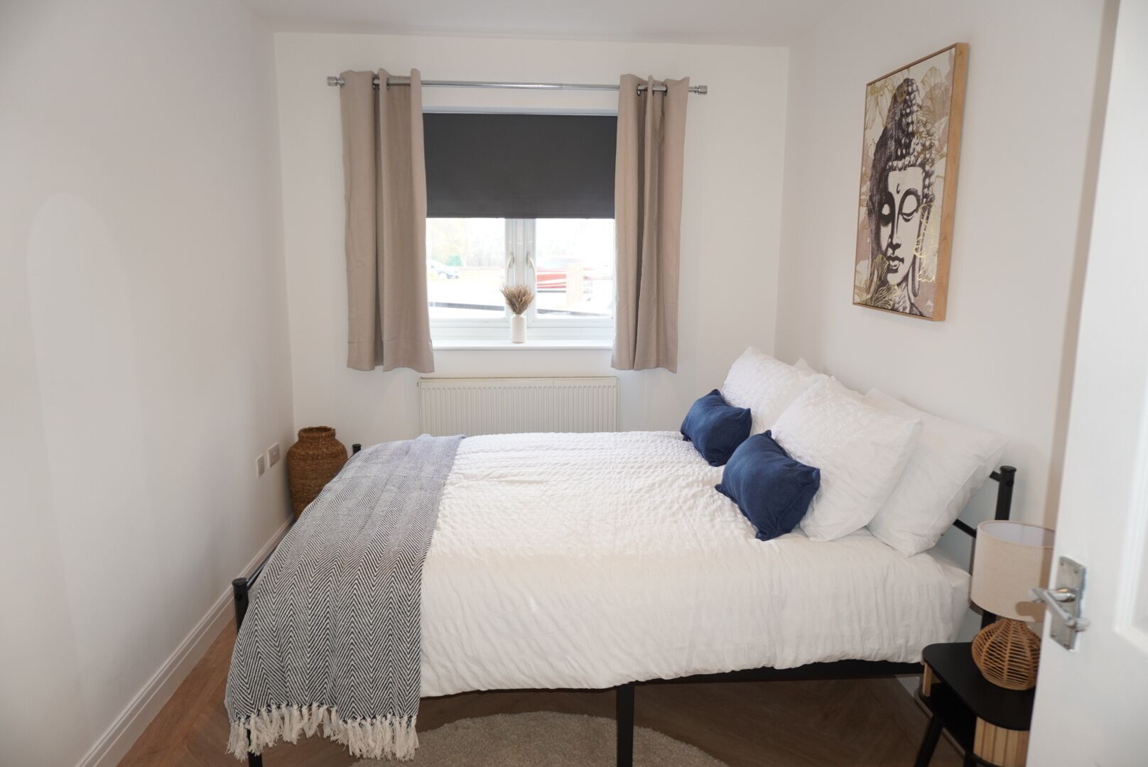 Un dormitorio ordenado con cama, ventana con cortinas, radiador y una obra de arte en la pared.