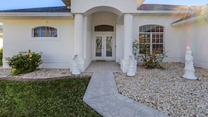 Una casa moderna con fachada blanca, una entrada central con puerta de vidrio y un pequeño jardín con estatuas blancas y un sendero.