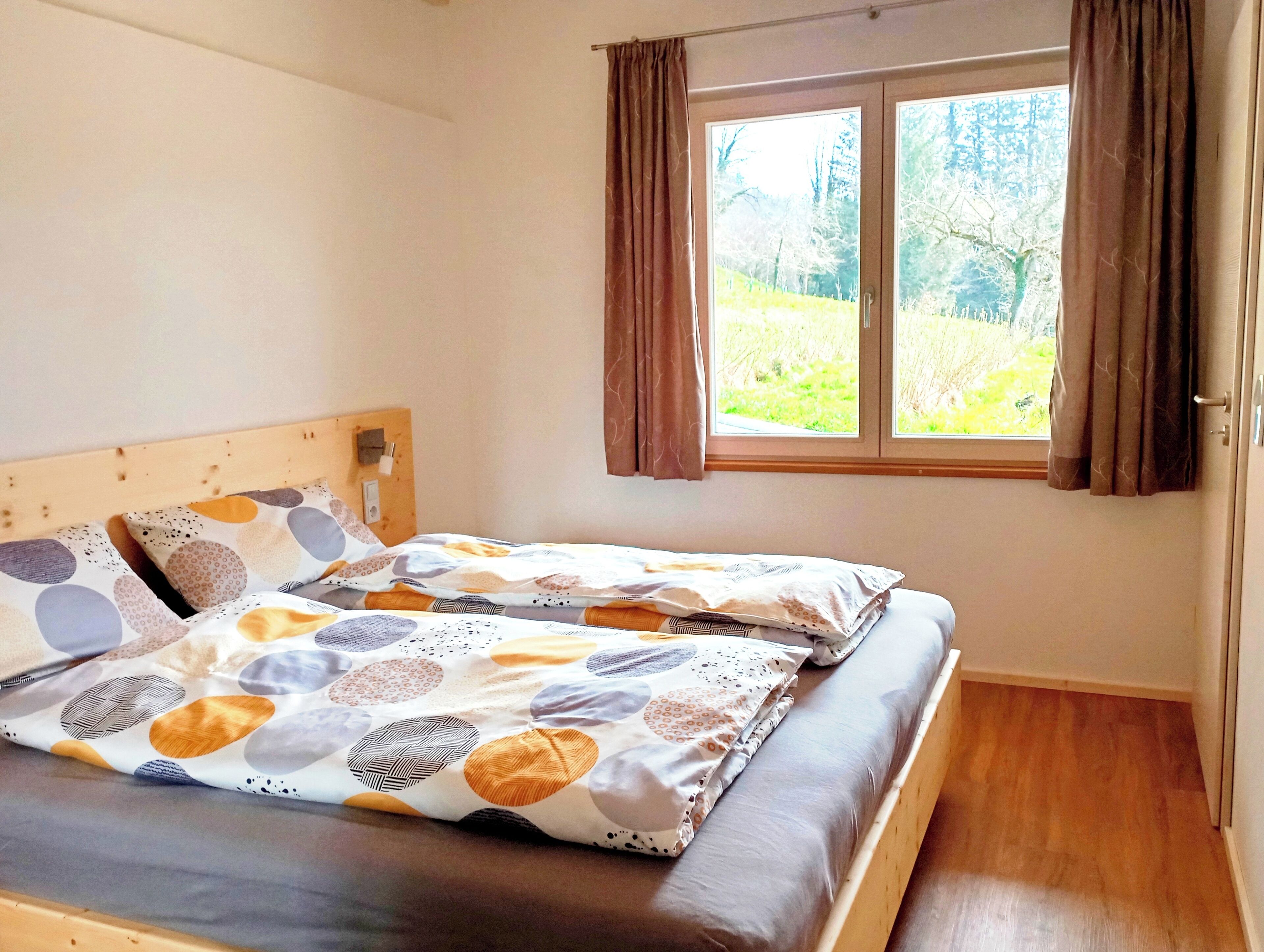 Une chambre à coucher avec une tête de lit en bois, un lit recouvert d’une literie à motifs, une fenêtre avec des rideaux et une vue sur la verdure à l’extérieur.