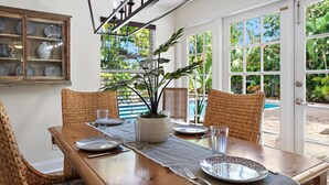 A dining area with a wooden table set for four, surrounded by wicker chairs. A potted plant is on the table, and there is a view of a pool and greenery outside. - Sawfish Bay Bungalow Old Florida Charm (Jupiter)