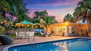A pool area with a lounge chair and umbrella, surrounded by palm trees and a wooden fence. - Sawfish Bay Bungalow Old Florida Charm (Jupiter)