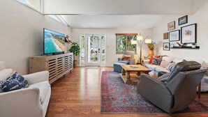 A spacious living room with a large TV, comfortable seating, a wooden cabinet, and a view through a glass door. - Sawfish Bay Bungalow Old Florida Charm (Jupiter)