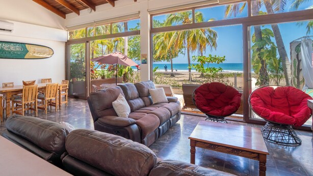 A spacious living area with a large window overlooking a beach, a wooden ceiling, and a dining area with wooden furniture. - Beachfront AC Villa, 3BR, 3.5BA,  great surfing! Free Parking (Guanacaste Province)
