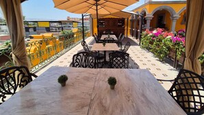 An outdoor patio with tables and chairs, a large umbrella, and a yellow building with arches in the background.