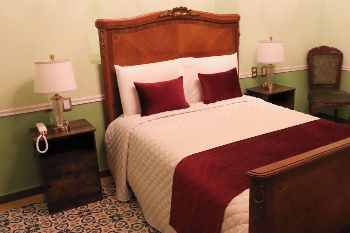 A neatly made bed with a wooden headboard, two bedside lamps, a telephone on a nightstand, and a chair in a room with patterned flooring.