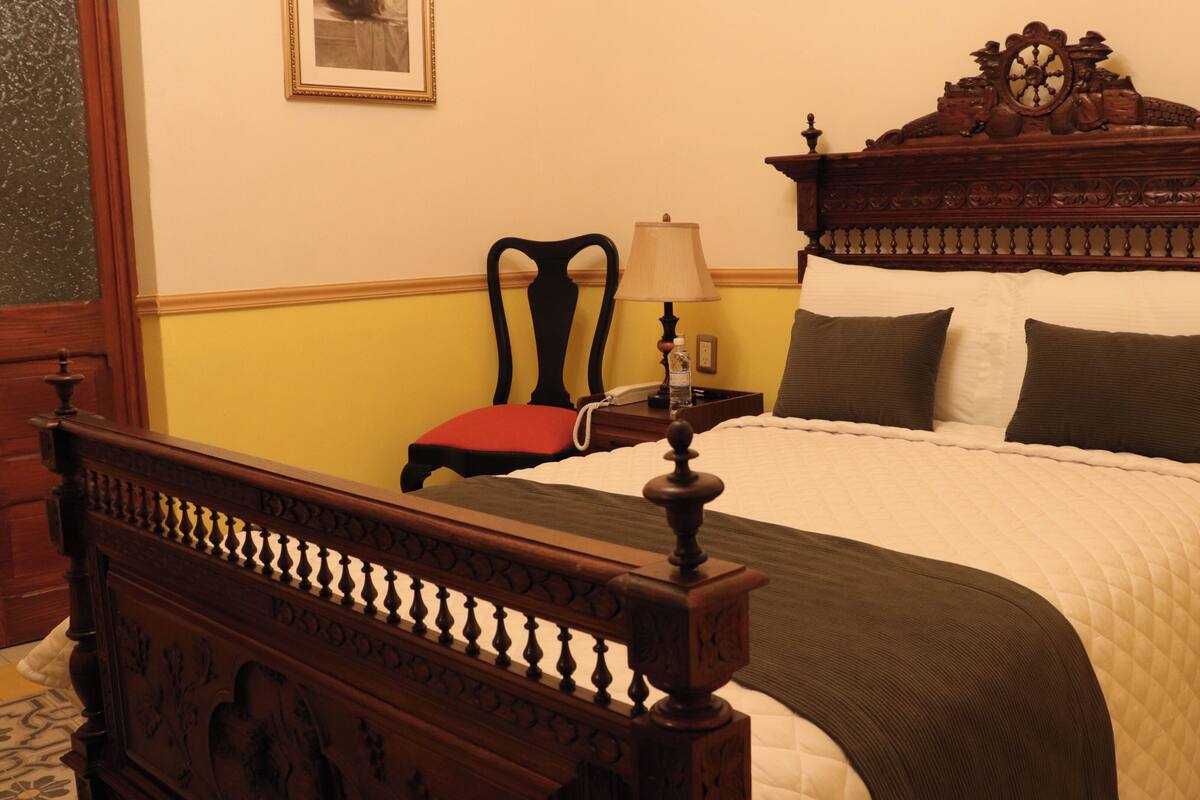 A bed with a wooden headboard, a bedside table with a lamp, a chair, and a framed picture on the wall.