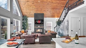 Una sala de estar moderna con chimenea, sofás de cuero, una zona de comedor con mesa de mármol y una encimera de cocina con comida y bebida.