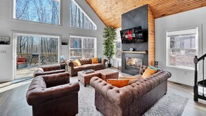Amplia sala de estar con ventanas grandes, chimenea y televisor montado.