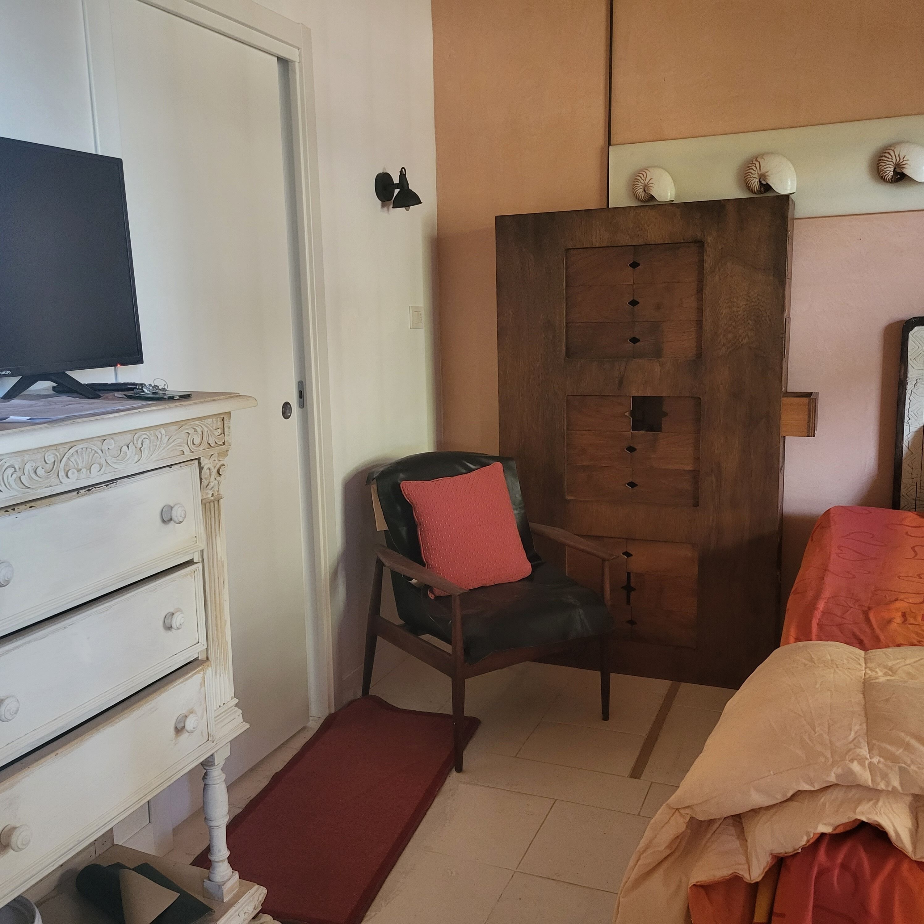 Ein Zimmer mit einem Bett, einem Holzschrank, einem Sessel mit rotem Kissen, einem Schreibtisch mit Computer und einem kleinen Teppich.