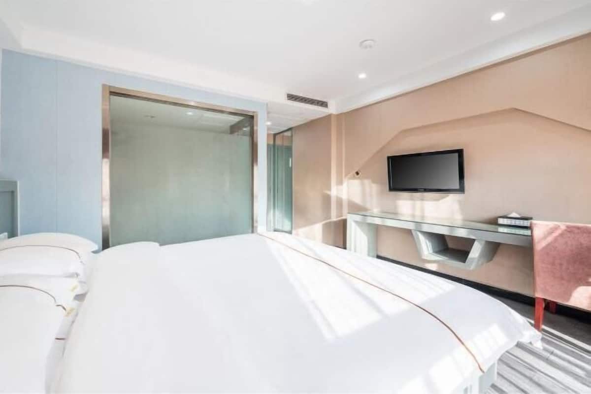 A modern hotel room with a large bed, a glass partition, a flat-screen TV, and a small table.
