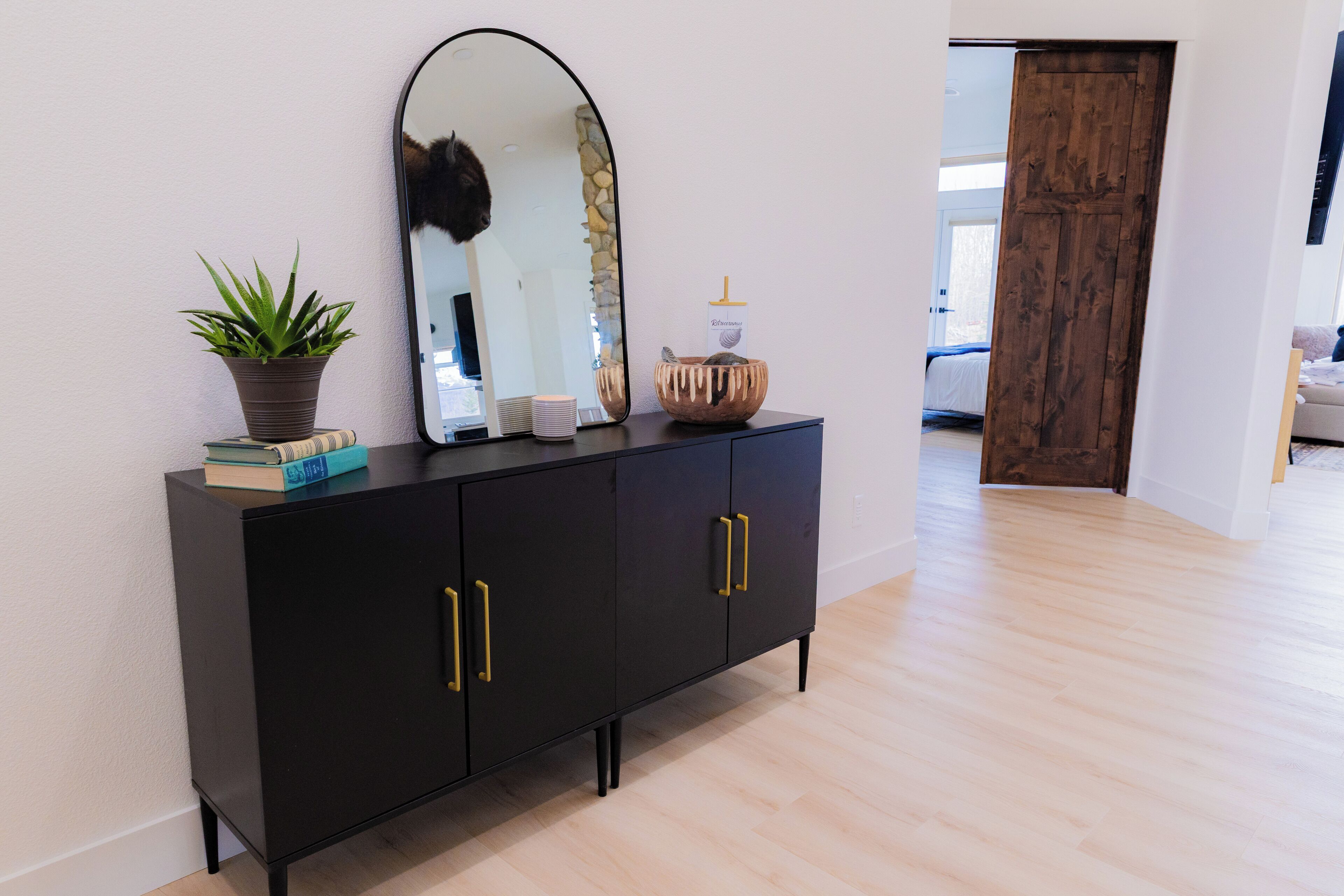A black cabinet with gold handles, a large mirror, a potted plant, and a decorative bowl.