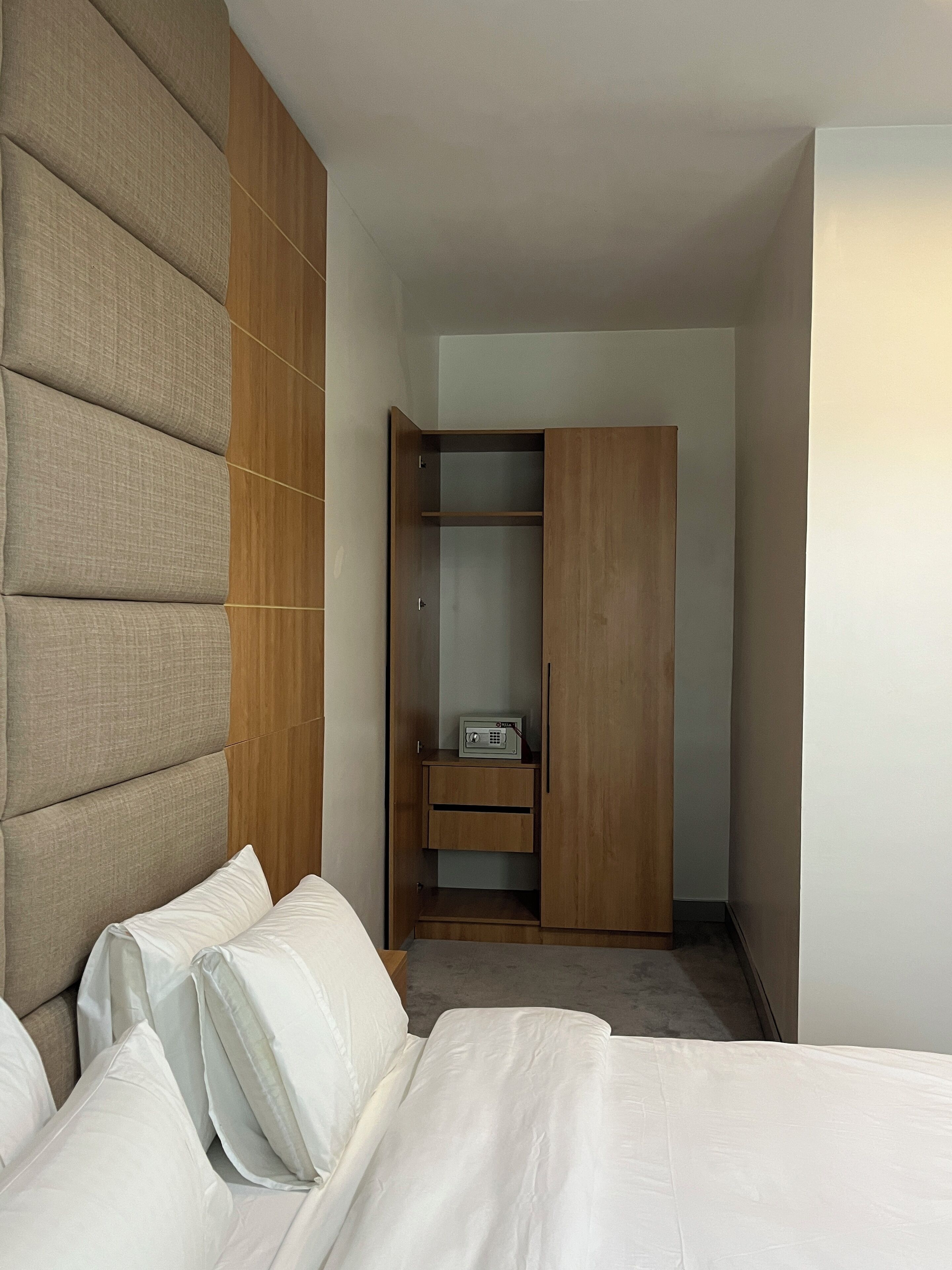 A hotel room with a bed, a large padded headboard, a wooden wardrobe, and a small bedside table with a radio.