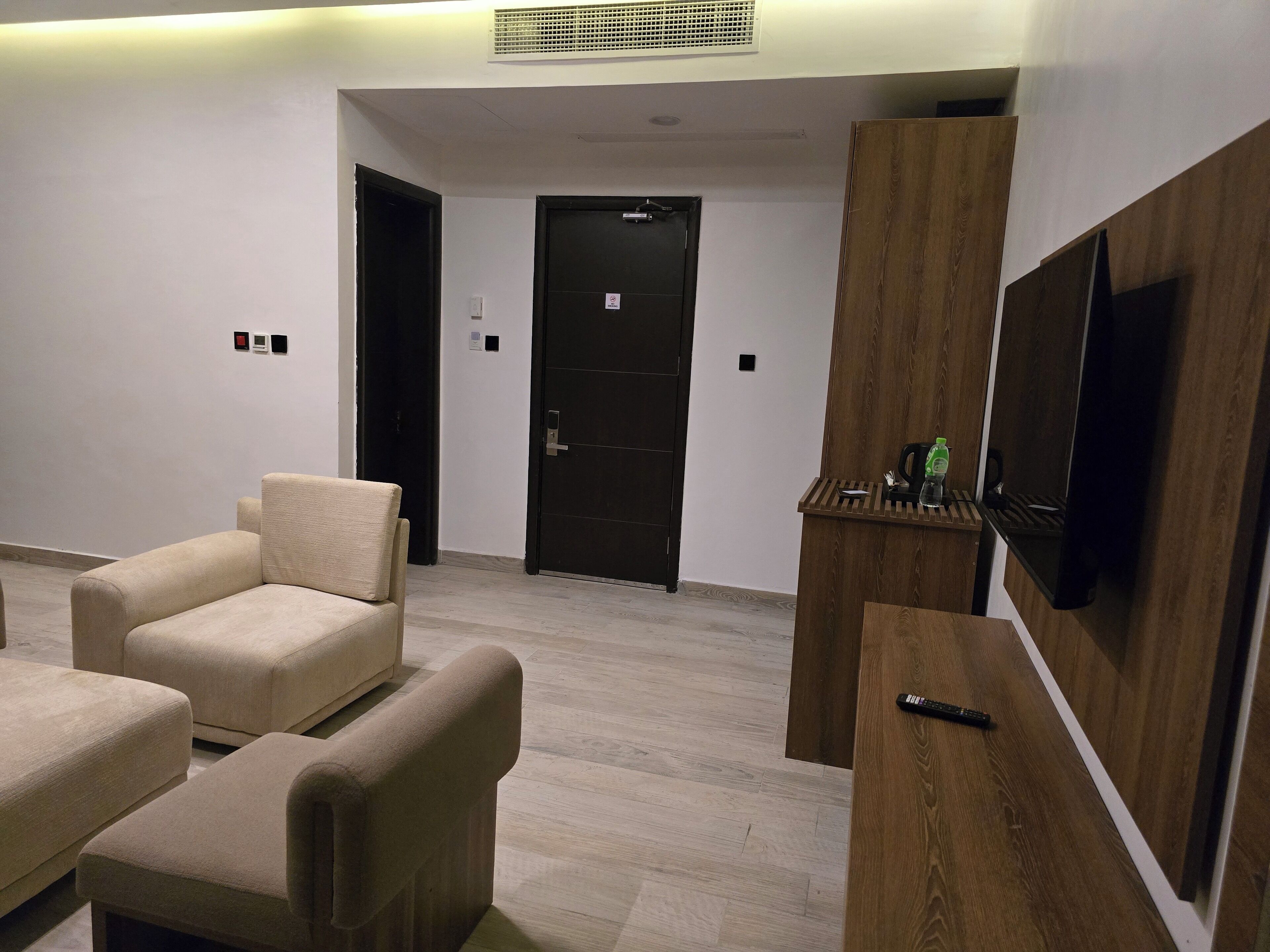 A modern living room with a flat-screen TV mounted on a wooden panel, beige armchairs, and a dark door in the background.