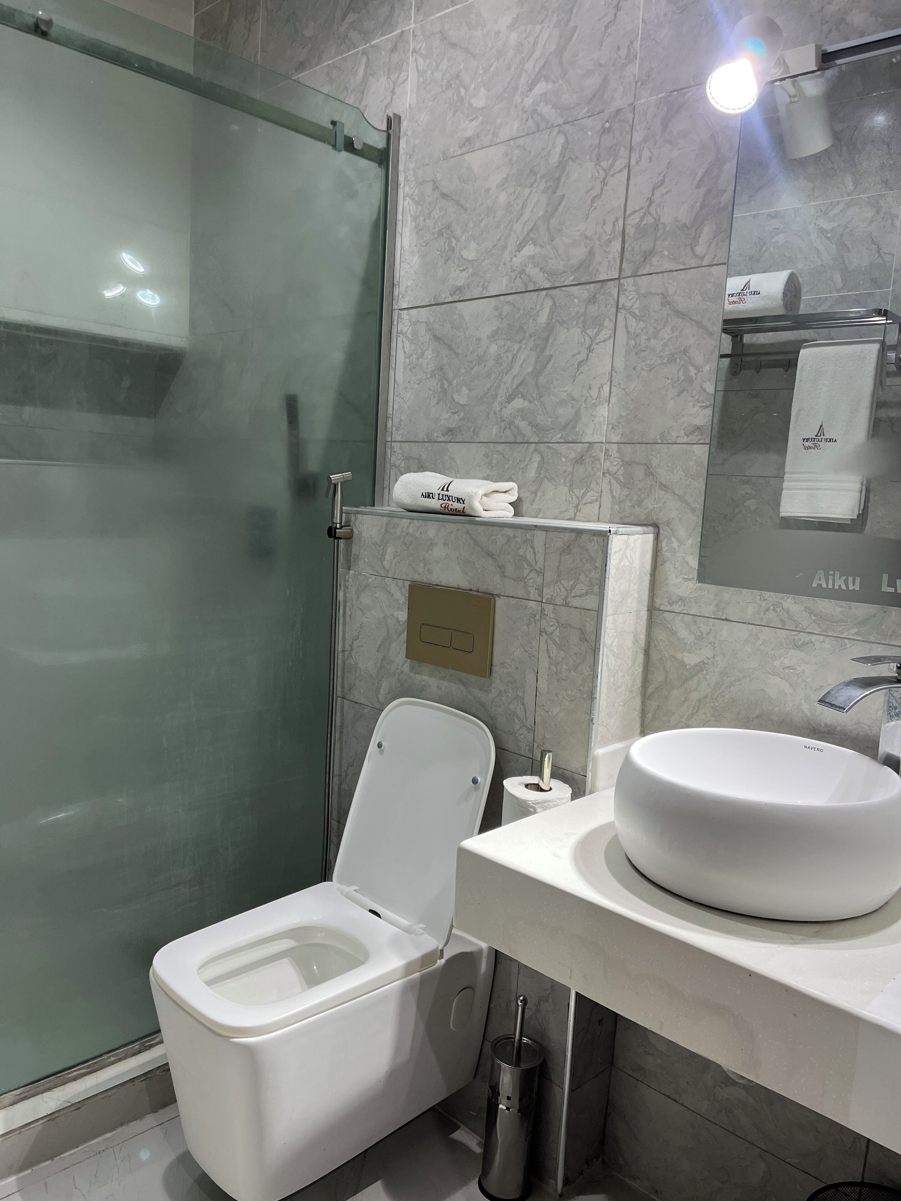 A modern bathroom with a glass shower enclosure, a white toilet, a round sink, and a mirror with a towel hanging on the wall.